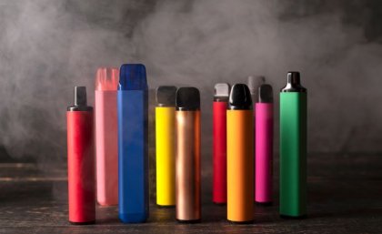 A row of coloured vapes stand upright on a wooden base, with vapour in the background.